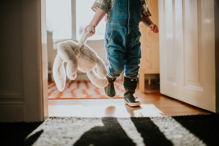A small child carrying a soft toy through an open front door